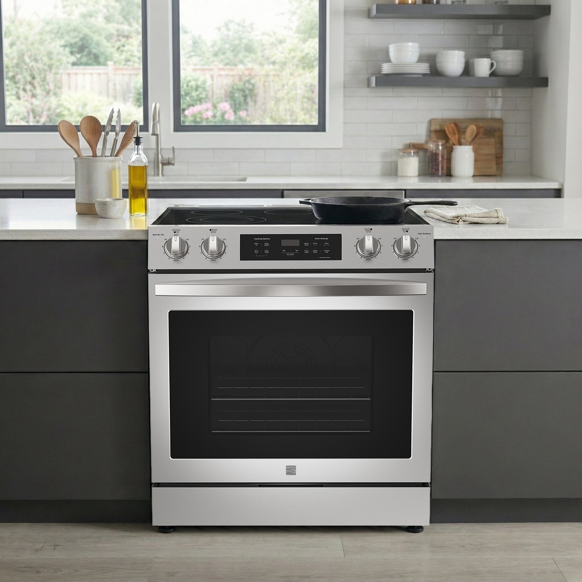 Kenmore stainless steel electric range with a cast-iron skillet on the stovetop installed in a bright, modern kitchen between dark grey cabinets