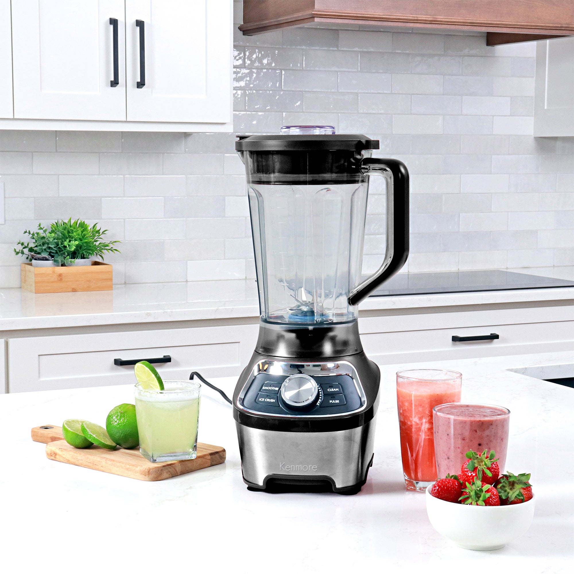 64 oz stand blender on a white countertop with glasses of juice, smoothie, and frozen lime margarita and a bowl of strawberries arranged around it.