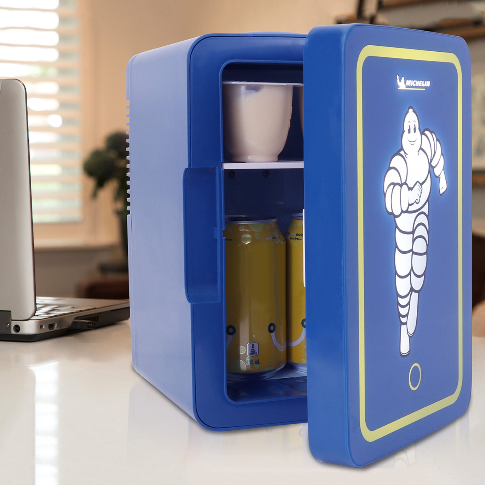Michelin mini fridge, partly open with cans of pop and a yogurt cup visible, beside a laptop computer on a white desktop